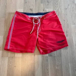 Aston Martin Racing x Hackett badshorts - Snygga röda badshorts från Hackett x Aston Martin Racing med vita kontrastsömmar, snörning i midjan och en bakficka med lock. Shortsen har sportig känsla med randiga detaljer längs sidorna och broderad logga framtill. Perfekta för stranden eller poolen.