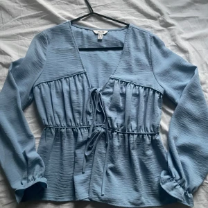 Blå blus - Supersöt ljusblå blus från Aware med v-ringning och knyt framtill. Blusen har långa ärmar med resår vid ärmslut och en lätt rynkad struktur som ger en chill vibe. Perfekt för dig som gillar romantiska detaljer och en luftig känsla.