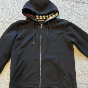 Svart Burberry Zip Hoodie - Svart hoodiejacka från Burberry med dragkedja och klassiskt rutigt foder i huvan. Jackan har broderad logga på bröstet, snörning i huvan och två stora fickor framtill. Tillverkad i mjuk bomull för en chill och stilren look.