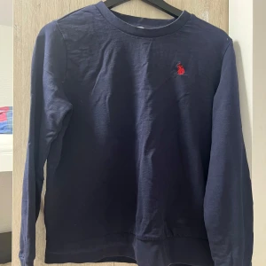 Mörkblå tröja från U.S. Polo Assn. - Snygg mörkblå långärmad tröja från U.S. Polo Assn. med klassisk rund halsringning och röd broderad logga på bröstet. Tillverkad i mjuk bomull som känns skön mot huden. Perfekt för dig som gillar stilren och enkel design. Aldrig använd