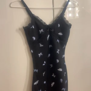 Svart kortklänning med spets och fjärilar🦋 - Supersöt svart kortklänning med smala axelband, spetsdetaljer vid urringningen och små blåvita fjärilar över hela tyget🤍 Klänningen har en figurnära passform och en liten rosett framtill. Perfekt för dig som gillar gulliga detaljer🙌🏼