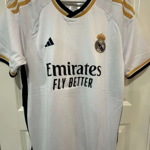 Real Madrid 2024/25 Bellingham 5  - Real Madrid matchtröja med Bellingham 5 på ryggen. Vit med guld- och svarta detaljer, klubbmärke broderat på bröstet och Adidas-logga. Korta ärmar, svart och guld på krage och ärmslut. Tillverkad i lätt och ventilerande material.