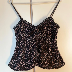 Blommig svart linnetopp från NA-KD - Supersöt svart linnetopp från NA-KD med små rosa och vita blommor. Toppen har smala axelband, spetsdetaljer vid urringningen och smockad rygg för extra komfort. Perfekt för varma dagar och ger en romantisk vibe till din outfit.