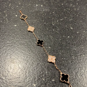 Armband med svarta och glittriga blommor - Snyggt armband med roséguldfärgad kedja och fem fyrklöverformade hängen. Tre hängen är svarta och två är glittriga med små stenar. Perfekt accessoar för att lyfta din outfit med en elegant touch.