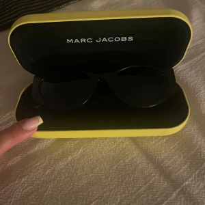 Svarta solglasögon Marc Jacobs - Snygga svarta solglasögon från Marc Jacobs med cool cat eye-form och breda skalmar med stor vit logga. Kommer i ett gult fodral. Perfekta för att ge din look en extra edge och skydda ögonen mot solen.