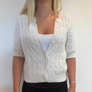 Vit kabelstickad kofta med kort ärm - Superfin vit kabelstickad kofta med korta ärmar och v-ringning. Koftan har ribbade muddar och knäpps framtill med vita knappar.