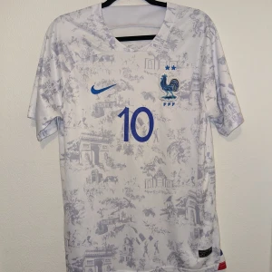 Nike Frankrike fotbollströja(kit) - Säljer en vit kortärmad fotbollströja med blå detaljer och nummer 10 på framsidan. Tröjan har ett unikt mönster och är märkt med en tupp och två stjärnor. Tillhörande blå shorts med nummer 10 och tuppemblem. Perfekt för fotbollsfans! Frankrike helkit med shorts och tröja. Kan diskuteras om pris 😊 Den är riktigt skön köpte den och använde den bara två gånger men annars i perfekt skick. 