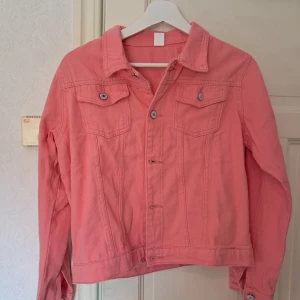 Rosa jacka i storlek M - Korallrosa jacka med klassisk krage, två bröstfickor med knappar och silverfärgade detaljer. Jackan är tillverkad i bomull och polyester, har långa ärmar och en rak passform. Perfekt för dig som vill sticka ut med färg i din outfit. Ganska liten i storlek