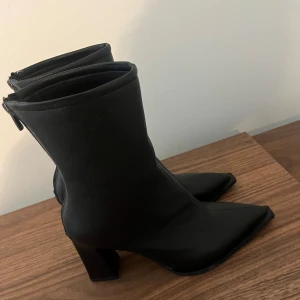 Svarta stövletter med klack och spetsig tå - Snygga svarta stövletter med spetsig tå och hög klack. Skorna har dragkedja bak och är gjorda i ett slätt, matt material som liknar skinn. Perfekta för dig som vill ha en edgy och stilren look. Helt nya