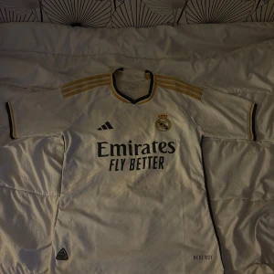 Real Madrid matchtröja Adidas vit/guld - Säljer en Real Madrid fotbollströja i nyskick med vita och guldiga detaljer samt svarta inslag. Säljer då den var lite för stor för mig. Tillverkad i lätt funktionsmaterial. Storlek M