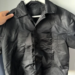 Prada skjorta kortärmad - Snygg svart skjorta från Prada med kort ärm och klassisk krage. Skjortan har en bröstficka med Pradas ikoniska metallogo och stängs med knappar framtill. Materialet är glansigt och ger en lyxig känsla, perfekt för dig som vill sticka ut med designerplagg.