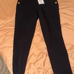 Svarta byxor med guldknappar XS - Snygga svarta byxor i storlek XS med hög midja och sex dekorativa guldknappar framtill. Byxorna har en slim passform och är tillverkade i ett stretchigt material som sitter tajt och bekvämt. Perfekta för dig som gillar en stilren look med lite edge.