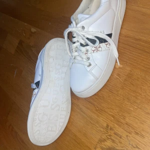 Vita skor från Guess - Snygga vita skor från Guess med klassisk logga på sidan och sula. Skorna har vita snören, rund tå och detaljer i brun,vitt och beige med mönster. Perfekta för en trendig och clean look.