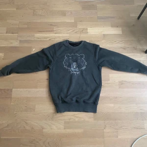 Mörkgrå Kenzo sweatshirt med tiger - Säljer en mörkgrå sweatshirt från Kenzo i storlek XS. Tröjan har ett stort broderat tigerhuvud på bröstet och klassisk rund hals. Mjuk och skön med ribbade muddar vid ärmslut och nederkant. Perfekt för dig som gillar streetstyle och vill sticka ut.