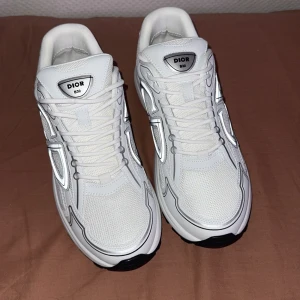 Vita Dior B30 sneakers storlek 43 - Säljer ett par fräscha vita Dior B30 sneakers med snygga silvriga detaljer och svart sula. Skorna har mesh och skinnpaneler, klassisk snörning och coola loggor både fram och bak. Perfekta för dig som vill sticka ut med lyxig streetstyle. Priset kan diskuteras;)