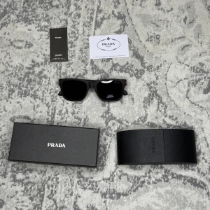 Svarta solglasögon från Prada - Snygga svarta solglasögon från Prada med breda bågar och polariserade linser. Klassisk fyrkantig form och röd detalj med Prada-logga på skalmen. Kommer med originaletui och certifikat. Perfekta för dig som vill ha en stilren och exklusiv look.