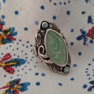 Vintageinspirerad silverring med grön sten - Unik ring i silverfärgad metall med en stor, oval grön sten i mitten och två små ljusblå stenar upptill och nedtill. Ringen har detaljerade snirkliga mönster runt stenen som ger en cool vintagekänsla. 