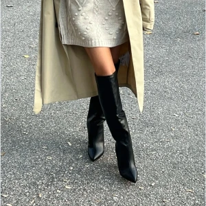 Svarta höga stövlar med klack - Snygga svarta stövlar i skinnimitation med spetsig tå och hög blockklack i storlek 36 från Nelly. Stövlarna går upp till knäet och har en stilren, minimalistisk look som passar perfekt till kjol eller klänning. Perfekta för dig som vill ha en trendig och edgy vibe. 