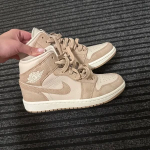 Beige Nike Air Jordan 1 High sneakers - Snygga Nike Air Jordan 1 High sneakers i beige och ljusbrun mocka och skinn i storlek 36. Klassisk siluett med snörning, perforerad tå och ikoniska Jordan Wings-loggan på sidan. Perfekta för dig som vill ha en clean och stilren look med streetkänsla.