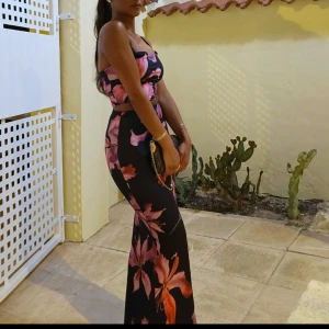 Svart långklänning med rosa blommor - Supersnygg svart långklänning med stora rosa och lila blommor. Klänningen har smala axelband och figurnära passform som framhäver siluetten. Materialet är mjukt och följsamt, perfekt för sommarkvällar. Blommönstret ger en färgstark och trendig vibe.