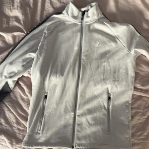 Vit fleecejacka Peak Performance L - Snygg vit fleecejacka från Peak Performance i storlek L. Jackan har hel dragkedja, höga fickor med dragkedja och diskret logga på bröstet. Perfekt för kyliga dagar och funkar lika bra till träning eller chill. Mjuk insida i fleece och sportig look.