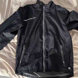 Svart vindjacka från Bauer, strl M - Svart vindjacka från Bauer med hel dragkedja och två fickor med dragkedja. Jackan har en sportig look, hög krage och meshfoder på insidan för extra komfort.