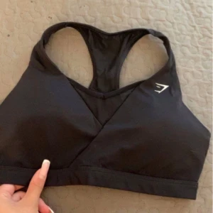 Svart sporttopp från Gymshark - Svart sporttopp från Gymshark med omlott-design framtill och brottarrygg. Toppen har en diskret vit logga på vänster sida och är tillverkad i ett stretchigt material som passar perfekt för träning.