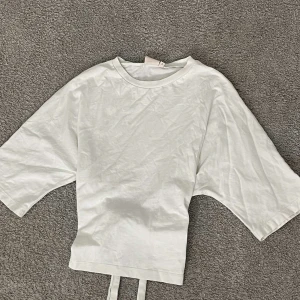 Vit omlottopp med knytband - Säljer en vit omlottopp med breda, korta ärmar och snygga knytband i ryggen. Toppen har en clean och enkel look med rund hals och croppad passform. Perfekt för dig som gillar minimalistisk stil och vill ha något som sticker ut med coola detaljer.