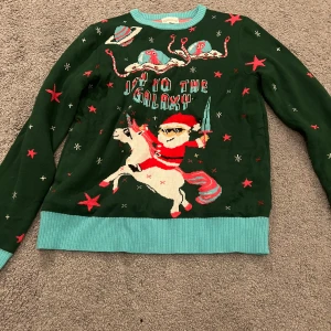 Grön jultröja med rymdtema från Christmas Sweats - Rolig grön stickad jultröja från Christmas Sweats med motiv av tomte som rider på en enhörning, rymdvarelser och texten 'Off to the Galaxy'. Tröjan har turkosa muddar och röda stjärnor över hela. Perfekt för dig som vill sticka ut på julfirandet!