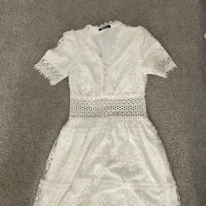 Vit spetsklänning med broderade detaljer - Superfin vit klänning från grina tricot med broderade spetsdetaljer och korta ärmar. V-ringad med dekorativ spetskant och markerad midja med hålmönster. Kjolen är lätt utställd och har transparenta spetsinslag. Perfekt för sommarens alla tillfällen!