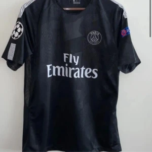 Svart PSG fotbollströja Adidas - Snygg svart PSG fotbollströja från Adidas med Fly Emirates-tryck på bröstet och Champions League-märke på ärmen. Tröjan har korta ärmar och diskreta geometriska mönster i tyget. Perfekt för dig som älskar fotboll och vill sticka ut på planen. Kan fixa custom nummer o namn det blir 85 kr dyrare 
