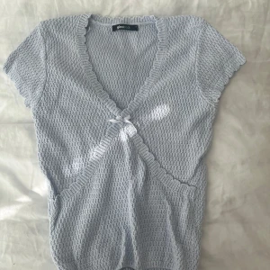 Ljusblå stickad omlottopp Gina Tricot - Supersöt ljusblå stickad topp från Gina Tricot med omlott-design och liten rosett framtill. Toppen har korta ärmar, v-ringning och volangkant längs ringningen. Perfekt för dig som gillar en gullig och trendig stil.