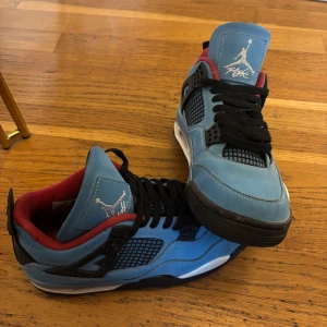 Nike Jordan 4 Cactus Jack blå sneakers - Säljer ett par Nike Jordan 4 Cactus Jack sneakers i blå mocka med svarta och röda detaljer. Skorna har svart snörning, vit och svart sula samt ikoniska Jumpman-loggan på plösen. Insidan är röd och meshpaneler på sidorna ger en sportig vibe.