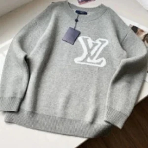 Grå stickad Louis Vuitton tröja - Säljer en grå stickad tröja från Louis Vuitton med stort vitt LV-logo framtill. Tröjan har rund hals, ribbade muddar och lång ärm. Perfekt för dig som gillar exklusiva märken och vill ha en clean look. Den passar S!