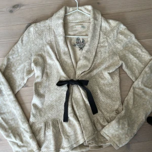 Beige omlottkofta från Odd Molly - Söt beige kofta från Odd Molly med diskret rutigt mönster, broderad logga på ryggen och svart knytband framtill.  Tröjan har volangkant nedtill och är långärmad, perfekt för lager-på-lager.