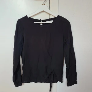 Svart blus med snörning från Cubus - Snygg svart blus från Cubus i storlek 36. Blusen har långa ärmar och rund halsringning, samt en cool snörningsdetalj längst ner framtill. Materialet känns lätt och luftigt, perfekt för dig som gillar stilrena plagg med en twist.
