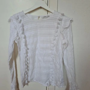 Vit blus med tofsdetaljer från KappAhl - Supersöt vit blus från KappAhl i storlek 38. Blusen har långärmade ärmar, snygga tofsdetaljer längs framsidan och diskreta ränder i tyget. Volang vid axlarna ger en extra touch. Perfekt för dig som gillar romantisk och feminin stil.