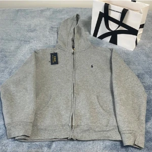 Grå🩶Ralph Lauren hoodie - Snygg grå🩶hoodie från Polo Ralph Lauren Tröjan har dragkedja, huva och två fickor framtill. Mjuk insida i fleece och klassisk Polo-logga broderad på bröstet. Perfekt för chill dagar🔥. 