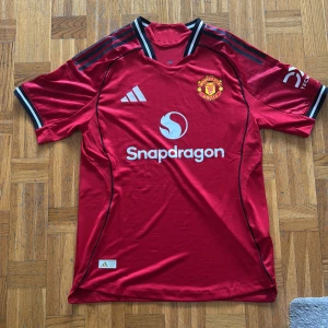 Manchester United röd matchtröja Adidas - Helt ny. Säljer nu vi en officiell Manchester United fotbollströja från Adidas i rött med svarta och vita detaljer på ärmarna. Tröjan har klubbmärket på bröstet, Snapdragon sponsortryck och klassiska Adidas-ränder på axlarna. Materialet är lätt och ventilerande polyester. Storlek XL men passar L.