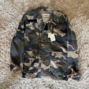 Camouflagejacka från Zadig & Voltaire - Snygg camouflagejacka från Zadig & Voltaire med klassisk krage, stora fickor framtill och dragkedja. Jackan är i bomull och har en loose passform, perfekt för en cool streetstyle-look. Färgerna går i grönt, beige och brunt.