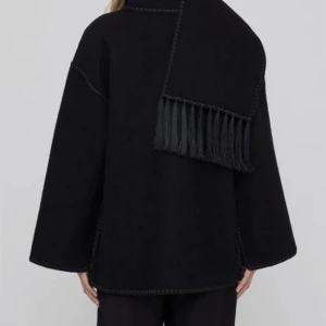 Storlek L - Svart oversized jacka i ull med breda ärmar och dekorativ fransdetalj över axeln. Jackan har synliga sömmar och en rak, avslappnad passform. Perfekt för dig som gillar minimalistisk stil med en twist.