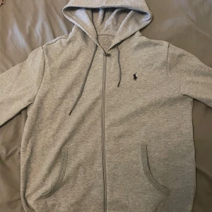 Grå hoodie från Polo Ralph Lauren - Snygg grå hoodie från Polo Ralph Lauren med dragkedja och huva. Klassisk logga broderad på bröstet, två fickor fram och snörning i huvan. Perfekt för chill dagar och enkel att matcha med jeans eller joggers. Mjuk bomullskänsla och långärmad modell.