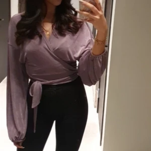 Lila omlottopp med knytband - Säljer en lila omlottopp med långa puffiga ärmar och breda knytband i midjan. Toppen har v-ringning och är i ett mjukt, lätt glansigt material som ger en chill men ändå snygg vibe. Perfekt att styla med höga jeans eller kjol.