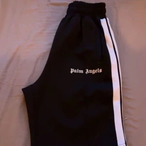 Palm Angels shorts - Svarta shorts från Palm Angels med vita sidoränder och elastisk midja. Klassisk logga broderad på vänster ben. Sportig och clean look, perfekta för en chill streetstyle. Materialet känns som polyester och passformen är loose.Från midja till längst ner på shortsen är det 56cm.