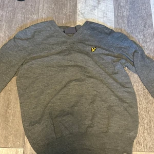 Grå v-ringad tröja från Lyle & Scott - Snygg grå v-ringad tröja från Lyle & Scott med den klassiska gula fågel-loggan broderad på bröstet. Tröjan är långärmad och tillverkad i mjukt material som passar perfekt till lager-på-lager-stil. Enkel att matcha med både jeans och chinos.