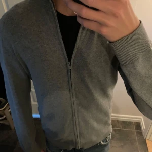 Grå kofta med dragkedja från Pier One - Snygg grå kofta från Pier One med hel dragkedja och ribbade muddar. Tröjan har hög krage och två fickor framtill. Perfekt för lager-på-lager och funkar året runt. Materialet är mjukt och stickat, vilket gör den både bekväm och stilren. Pris kan diskuteras!!!