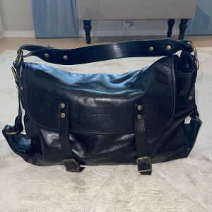 Svart axelväska i skinn från ULRIKA - Snygg svart axelväska i mjukt skinn från ULRIKA med justerbar rem, mässingsfärgade detaljer och spännen framtill. Väskan har lock med två remmar, dragkedjeficka på baksidan och rymligt huvudfack. Perfekt för dig som vill ha en stilren och praktisk väska. Ordpris 3495kr 