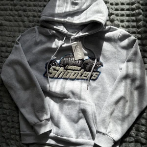 Grå Trapstar London Shooters  - Snygg grå hoodie från Trapstar med broderad London Shooters-logga i svart, grått och blått framtill. Klassisk känguruficka, snörning i huvan och mjukt material. Perfekt för dig som gillar streetwear och vill sticka ut med en fet logga. Storlek S men passar till M också 