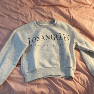 Grå sweatshirt med Los Angeles-tryck - Säljer en ljusgrå sweatshirt med stort svart Los Angeles-tryck på bröstet. Tröjan har rund hals, ribbade muddar vid ärmslut och nederkant samt lång ärm. Perfekt för dig som gillar streetstyle och enkel design.