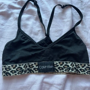 Svart linnetopp från Calvin Klein - Svart linnetopp från Calvin Klein med smala justerbara axelband och bred resår nertill i leopardmönster. Toppens resår har tydlig logga framtill och ger en cool kontrast till den enkla svarta designen. Perfekt för dig som gillar streetstyle och djurmönster.